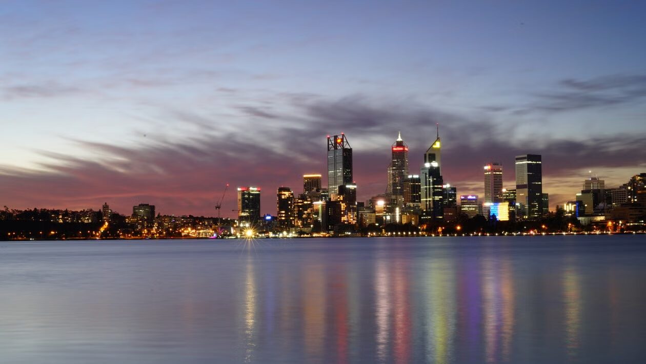 Perth skyline at sunset reflecting in the water, showcasing the vibrant city lights and evening sky hues.